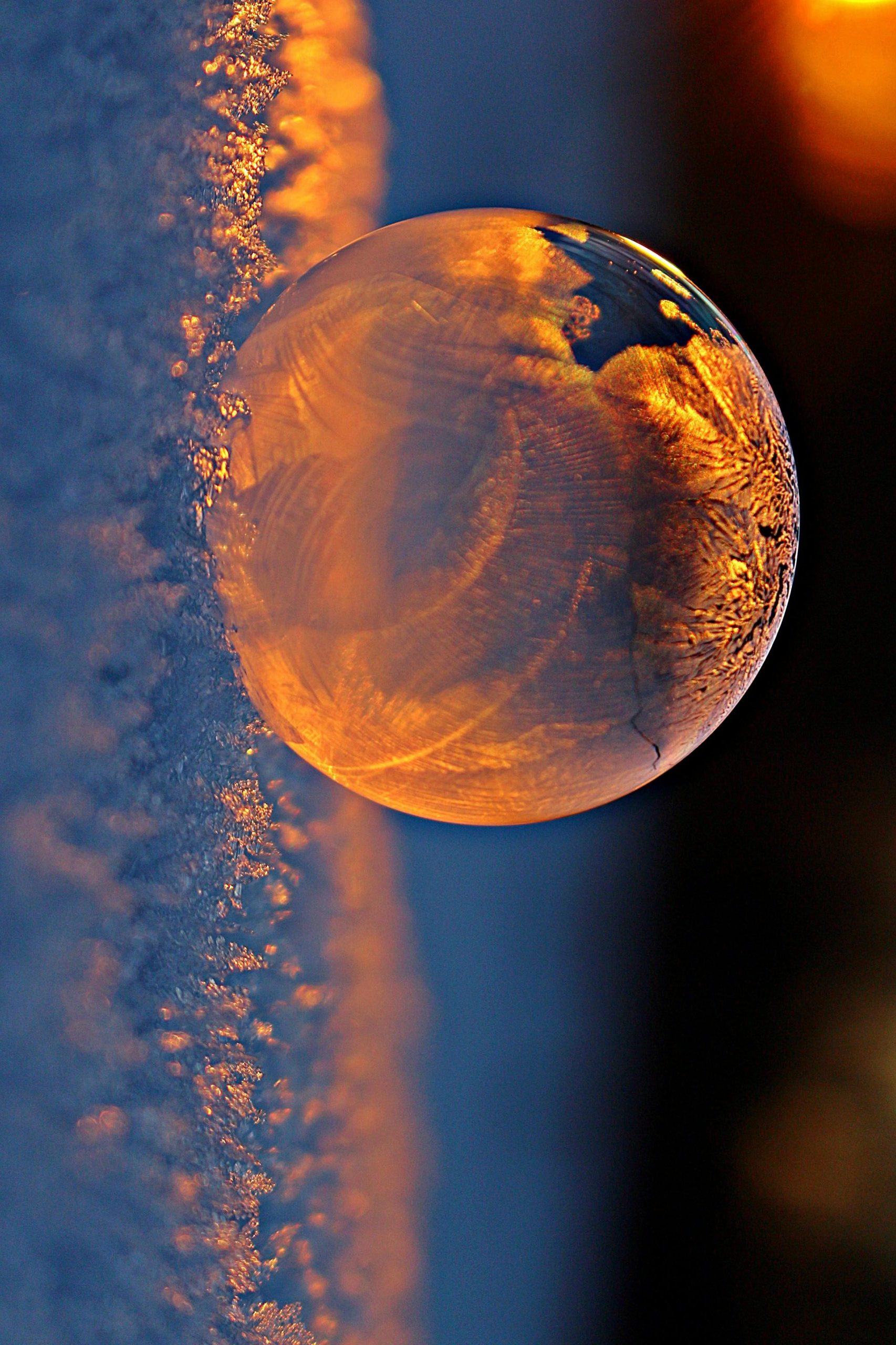 Close-up shot of a frozen bubble with warm reflections resting on a snowy surface at twilight.